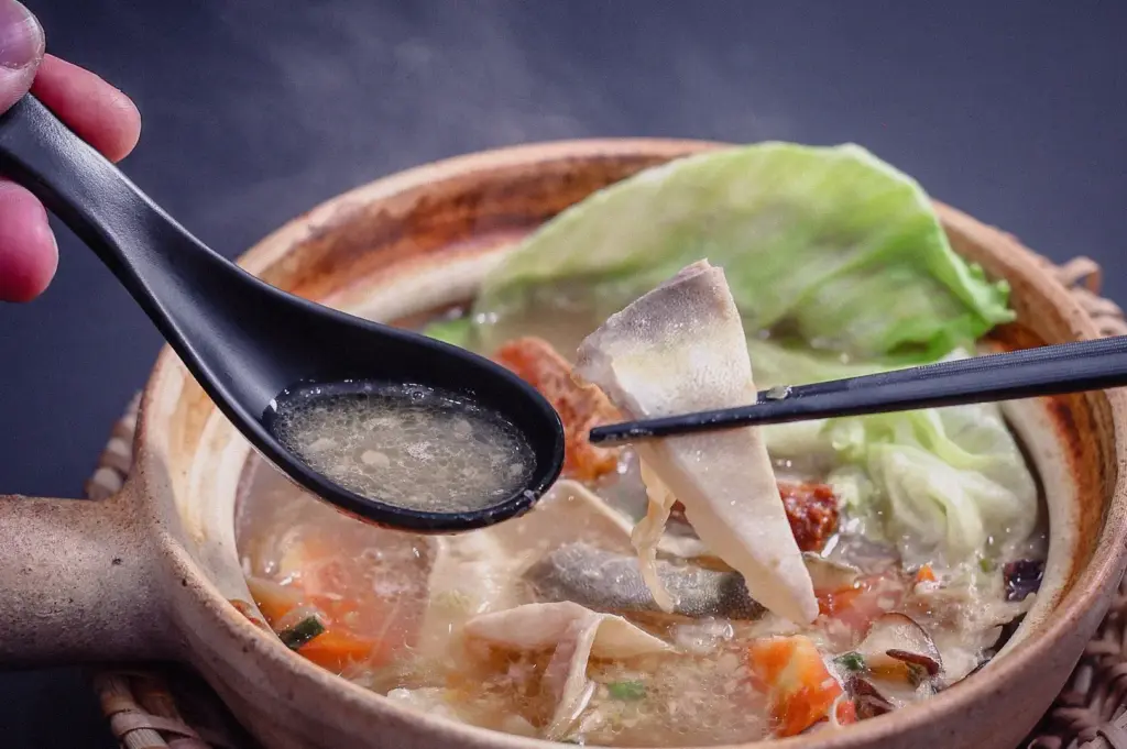 Eye-level close-up shot of Hup Lok's fish head steamboat in Singapore, featuring a spoon lifting collagen-rich six-hour pomfret broth while chopsticks hold fresh fish slices above a traditional claypot filled with seafood, cabbage, mushrooms, and vegetables.