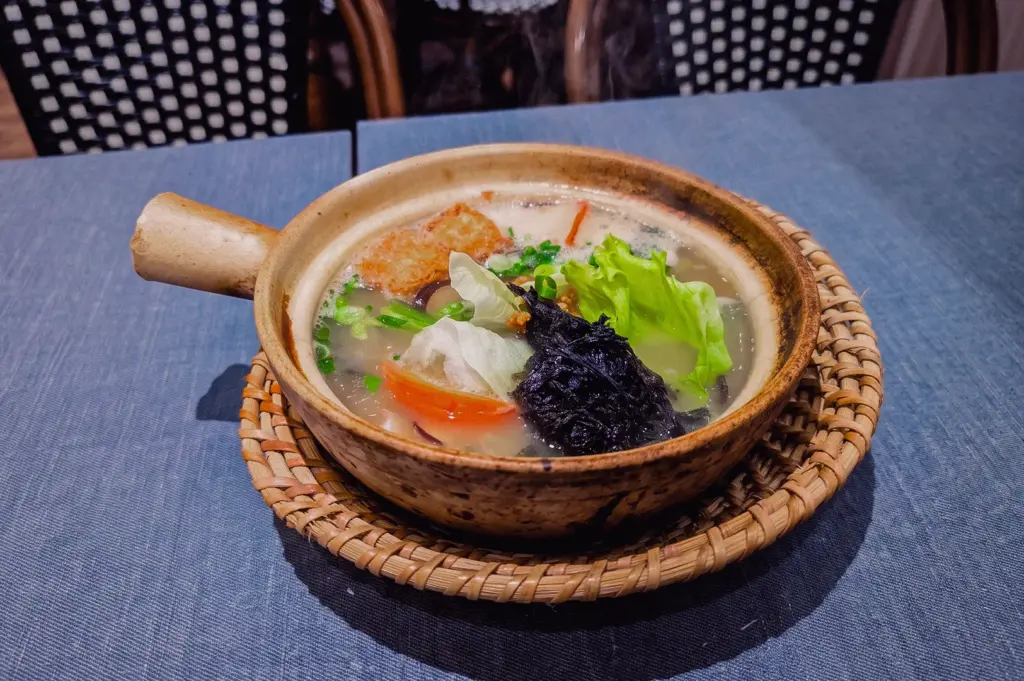 Traditional Singapore fish soup served in a rustic claypot with pomfret, vegetables, seaweed and rich collagen broth, photographed at a three-quarter angle in a restaurant setting.