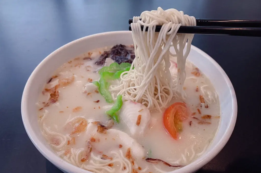 verhead close-up of creamy batang fish broth noodle soup, with thin noodles lifted by black chopsticks, surrounded by tomato slices, leafy greens, and seaweed, highlighting the collagen-rich texture that pairs well with noodles or bee hoon.