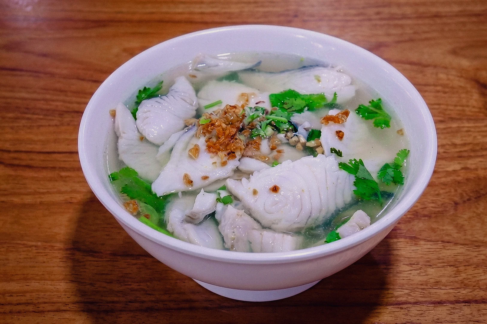 : High-angle shot of a white ceramic bowl filled with Teochew-style clear fish soup, loaded with tender sliced fish fillets, topped with aromatic fried garlic, crispy shallots, and fresh cilantro, resting on a dark wooden dining table.