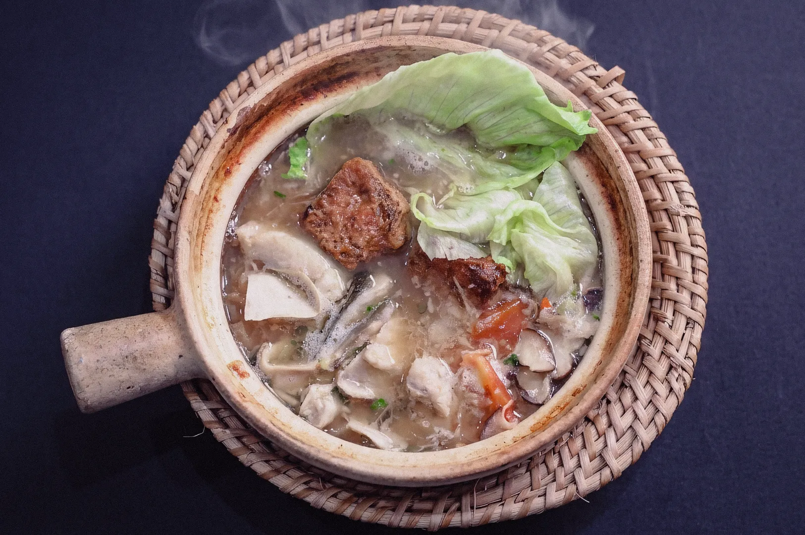 Steaming clay pot fish soup shot from a 45‑degree angle, showcasing rich collagen broth with lettuce, tofu, mushrooms, tomato slices, and braised meat chunks. Rustic woven mat base highlights traditional Asian comfort food, evoking Singapore fish soup culture and hearty home‑style dining.