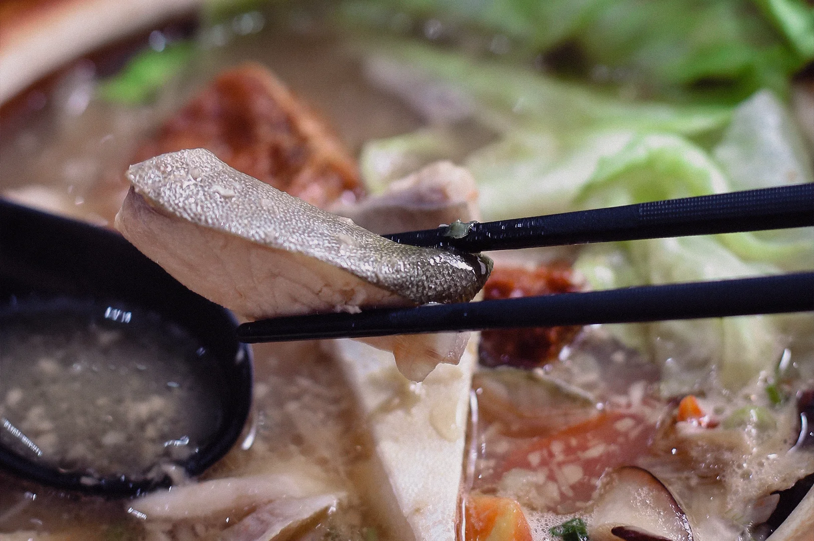 Close-up shot view of traditional restaurant fish soup served in a clay pot with pomfret fish on chopstick, tofu, seaweed, lettuce and collagen-rich fish broth, showcasing a premium Singapore fish soup dining experience.