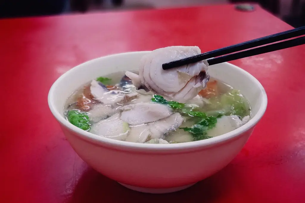 High-angle shot of dark chopsticks lifting a thick, tender slice of fresh fish fillet out of a steaming bowl of clear Singapore-style sliced fish soup resting on a red table.
