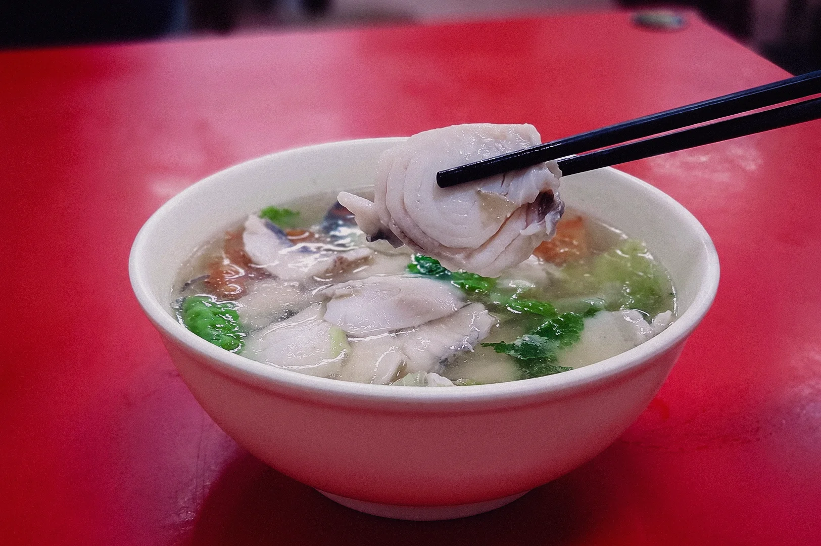 High-angle shot of dark chopsticks lifting a thick, tender slice of fresh fish fillet out of a steaming bowl of clear Singapore-style sliced fish soup resting on a red table.