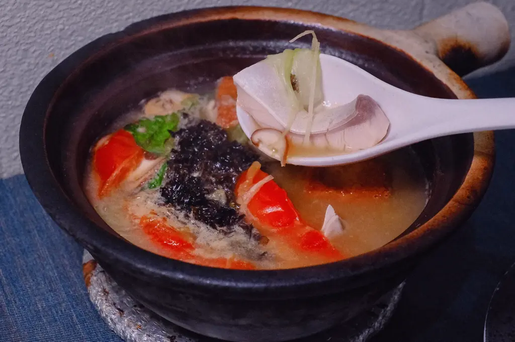 Close-up 45-degree angle of Grandpa's Pomfret Fish Soup featuring tender pomfret fish, shiitake mushrooms, ginger, tomato, and collagen-rich fish broth in a traditional claypot at a Singapore restaurant.