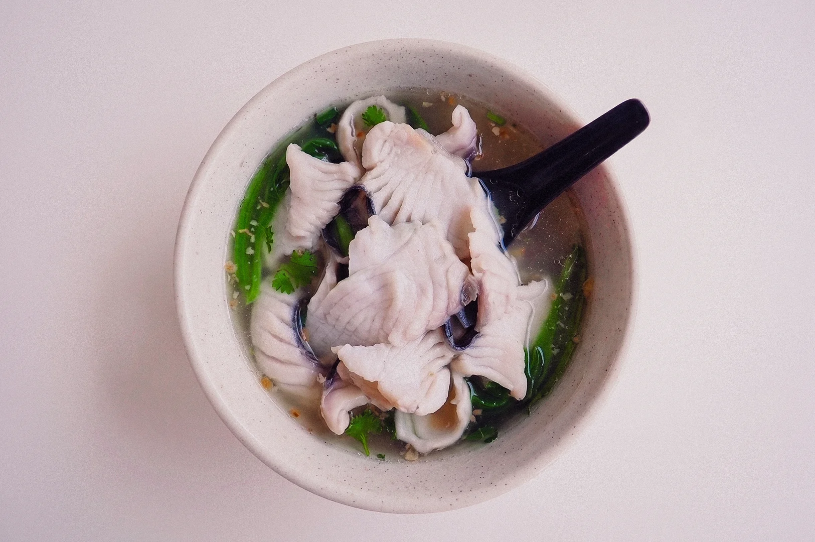 Top-down, overhead flat-lay shot of a light ceramic bowl filled with Singapore Teochew-style clear fish soup, packed with fresh sliced fish fillets, green vegetables, and garnishes, with a black soup spoon resting inside.