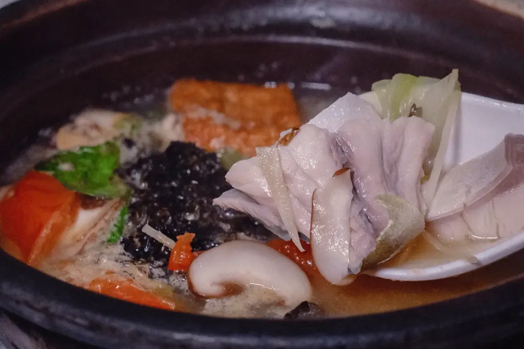 Close-up high-angle shot of a spoon scooping tender, fresh pomfret fish slices over a piping hot claypot fish soup filled with mushrooms, seaweed, and tomatoes.