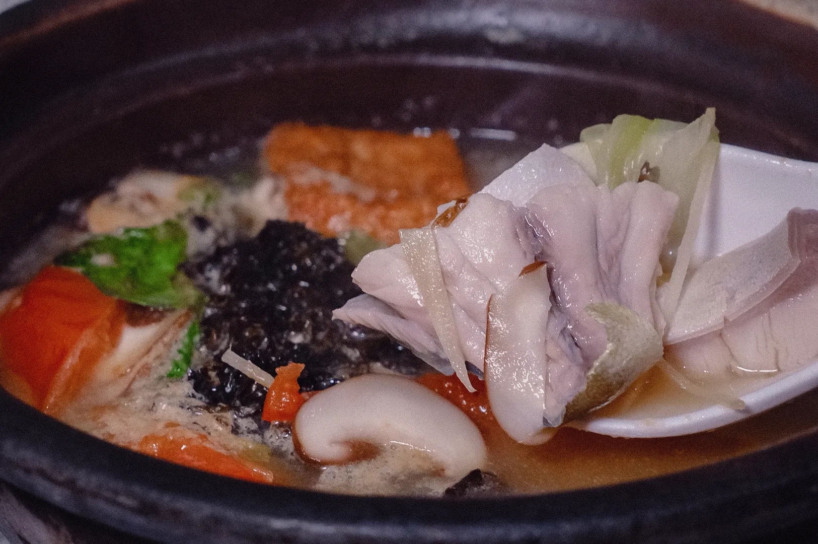 Close-up high-angle shot of a spoon scooping tender, fresh pomfret fish slices over a piping hot claypot fish soup filled with mushrooms, seaweed, and tomatoes.