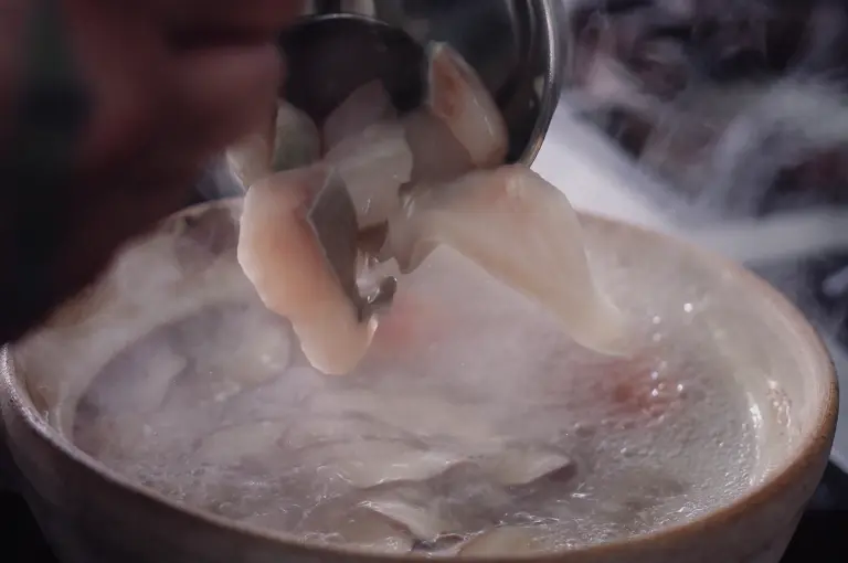Close-up shot of a metal ladle transferring delicate, raw pomfret slices into a steaming claypot filled with bubbling collagen broth.