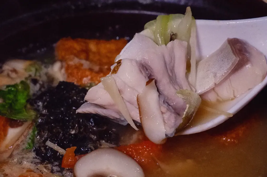 Close-up macro shot of tender pomfret fish slices lifted from a balanced fish soup with collagen-rich broth, shiitake mushrooms, seaweed and vegetables, photographed at an eye-level angle.