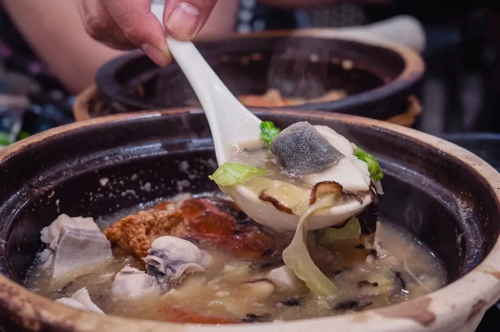 Eye-level close-up shot of a white soup spoon scooping tender pomfret fish, greens, and mushrooms out of a hot claypot broth.