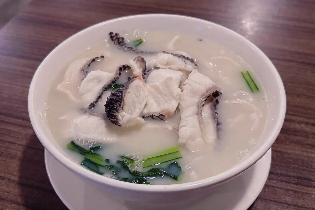 High-angle close-up shot of a white ceramic bowl filled with rich, opaque collagen-thickened milky fish soup generously topped with thick slices of fresh fish with dark skin edges, leafy green vegetables, and scallions, resting on a dark wooden table.