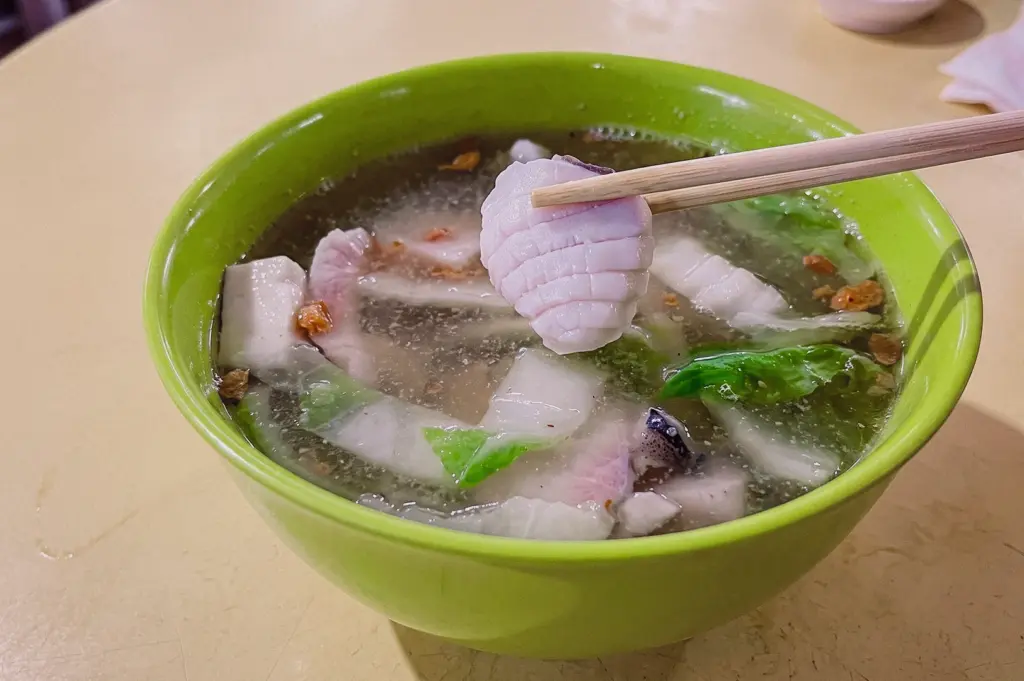 Close-up angled shot of fish soup with thick fish slices lifted by chopsticks above a bowl of clear broth, vegetables, and seafood, highlighting fish-to-broth ratio and portion quality.
