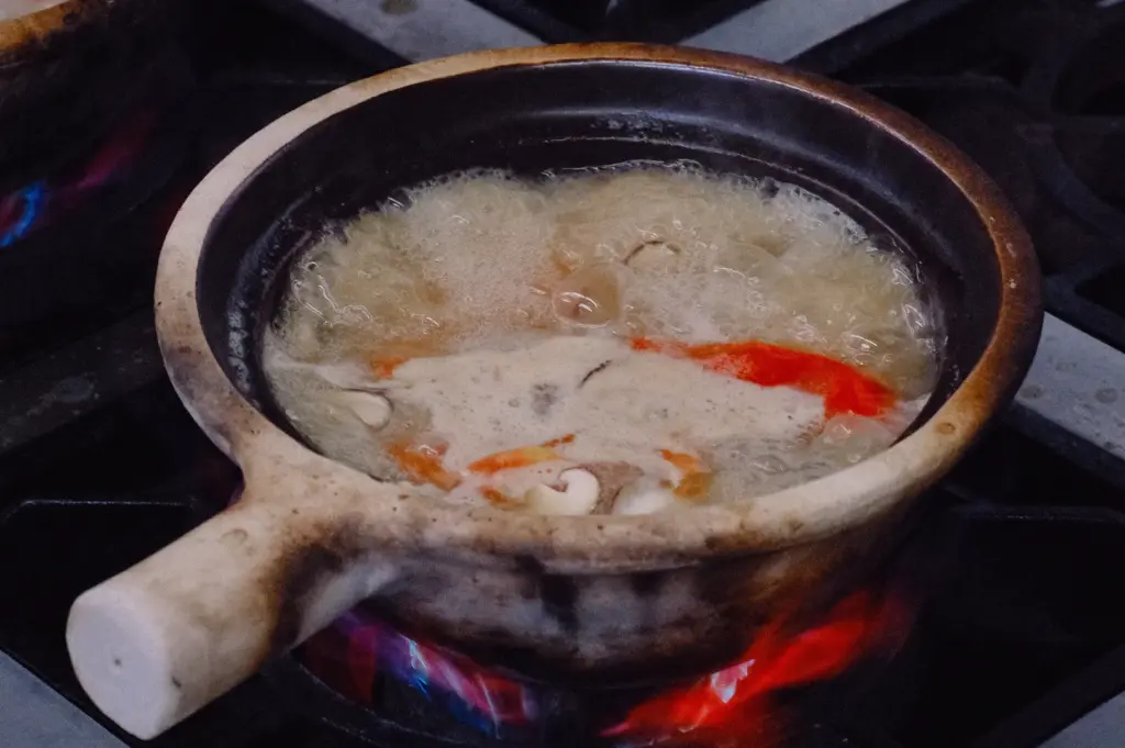 Side angle view of traditional clay pot fish soup boiling vigorously over a stovetop flame, with visible red tomato, seafood pieces, and vegetables in a collagen-thickened broth, capturing the rustic cooking process that enhances flavor and suits rice or thick bee hoon.
