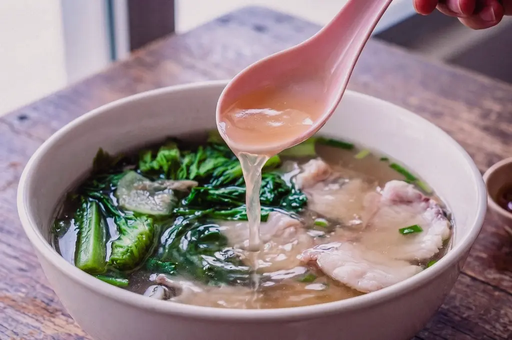 Close-up, high-angle shot of a pink soup spoon lifting clear, rich fish soup broth out of a deep porcelain bowl containing delicate fresh fish slices and vibrant green leafy vegetables.