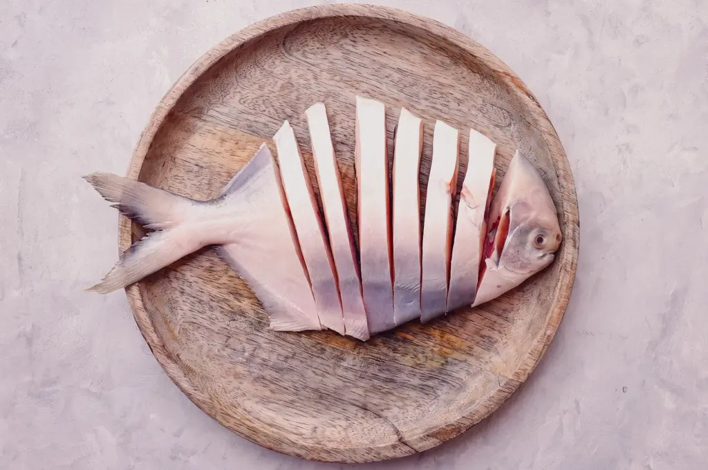 Top-down flat lay of freshly sliced pomfret fish on a wooden plate, showcasing premium fish preparation for traditional Singapore fish soup and seafood dishes.