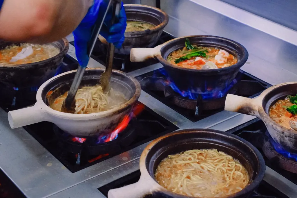 Fresh fish soup and noodles being prepared over open flames in a Singapore kitchen, captured from a high-angle action shot showcasing traditional cooking methods and collagen-rich fish broth preparation.