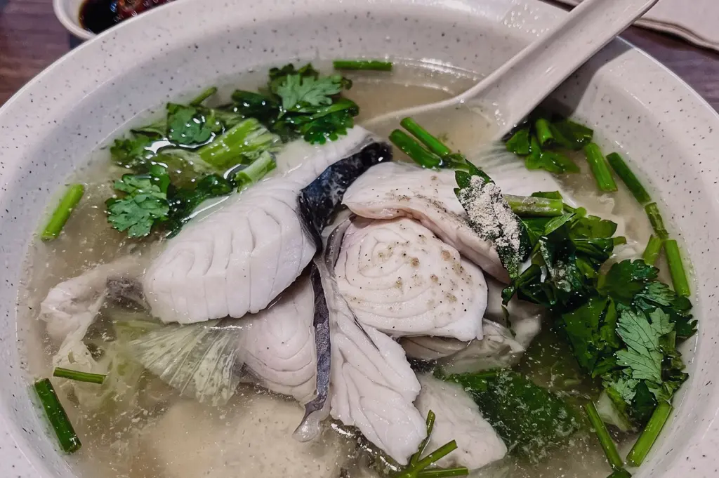 High-angle close-up of Batang Fish Soup featuring firm batang fish slices, fresh leafy greens and collagen-rich clear broth in a traditional Singapore fish soup bowl.