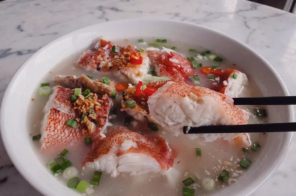 Close-up 45-degree angle of Grouper Fish Soup with chopsticks lifting a tender grouper fish slice from a clear collagen broth, garnished with spring onions, garlic and red chili in a Singapore seafood soup.