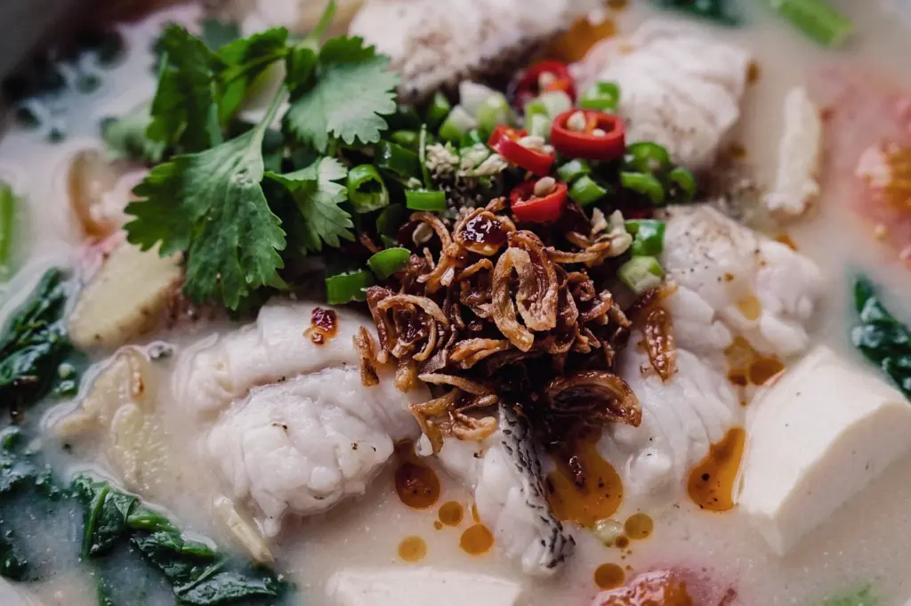 Close-up macro shot of Hup Lok's Grandpa's Pomfret Fish Soup featuring tender pomfret fish slices in collagen-rich broth, topped with crispy shallots, cilantro, spring onions, red chili, ginger, and tofu in a traditional Singapore fish soup.