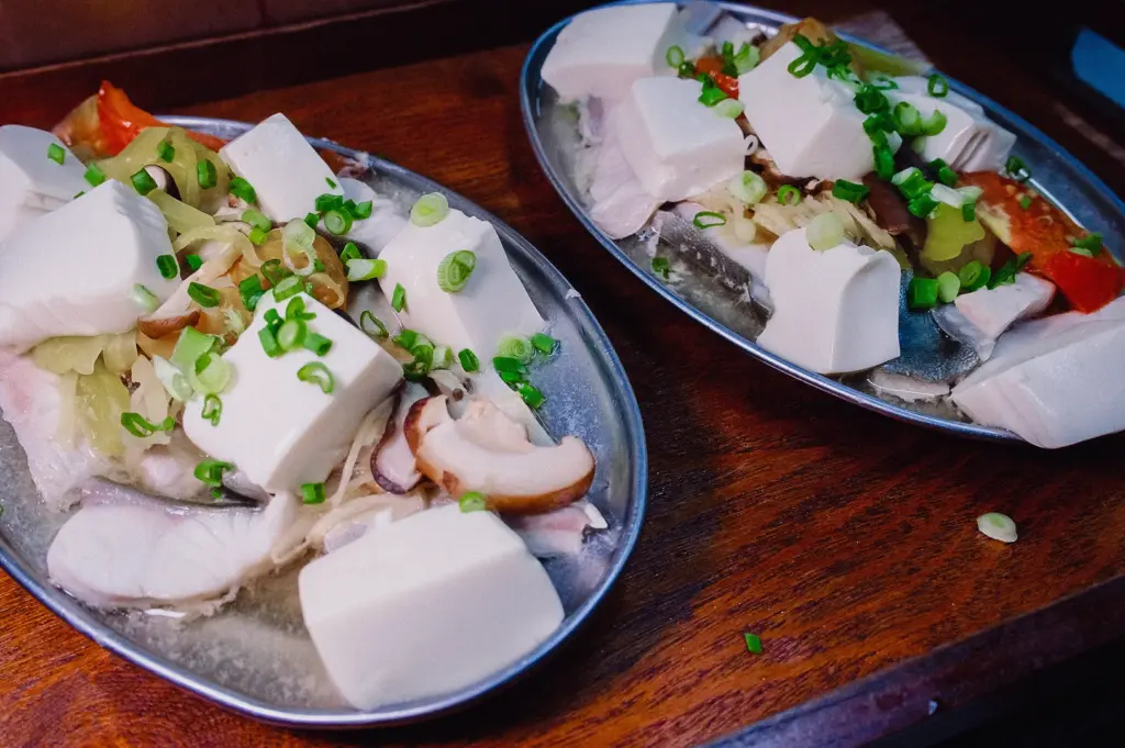 High-angle close-up shot of two oval metal platters laden with fresh pomfret slices, soft tofu cubes, shiitake mushrooms, and scallions prepared for Teochew steaming.
