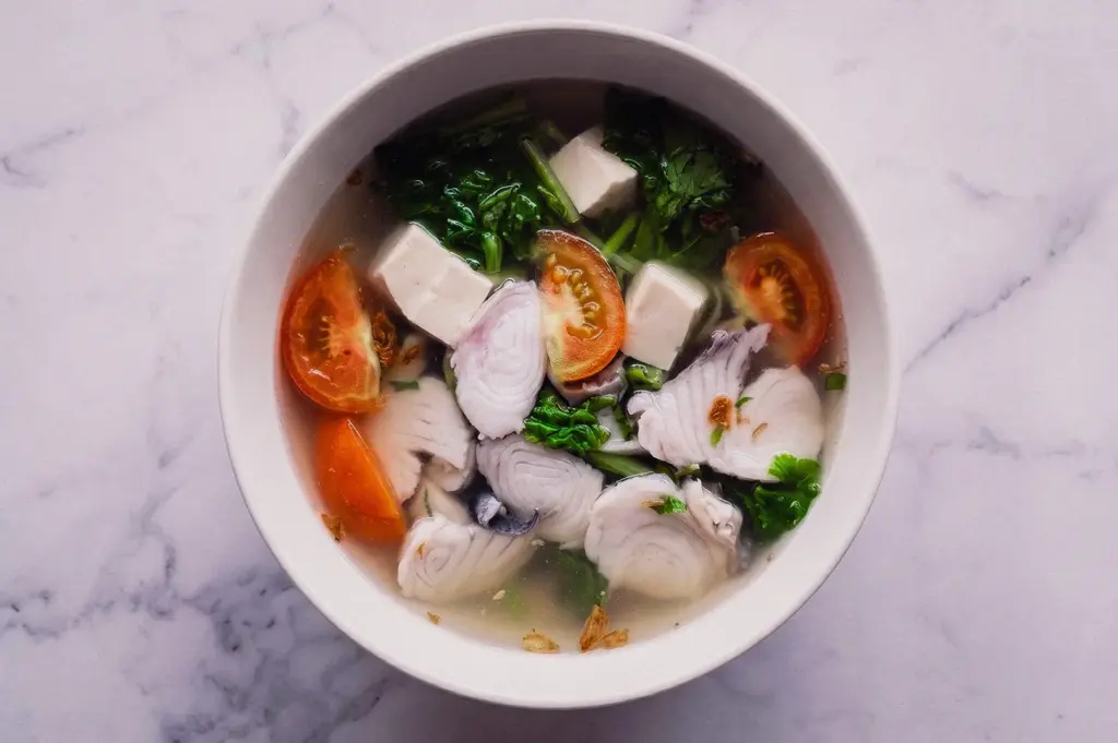 Top-down, overhead flat-lay shot of a classic bowl of Teochew fish soup featuring clean clear broth, sliced raw fish, fresh leafy greens, silken tofu blocks, sliced tomatoes, and cilantro served on a white marble surface.