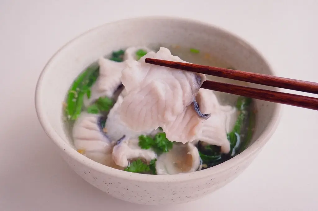 Close-up, eye-level shot of wooden chopsticks lifting a tender, freshly cooked slice of fish fillet out of a steaming bowl of fish soup, highlighting its firm texture.