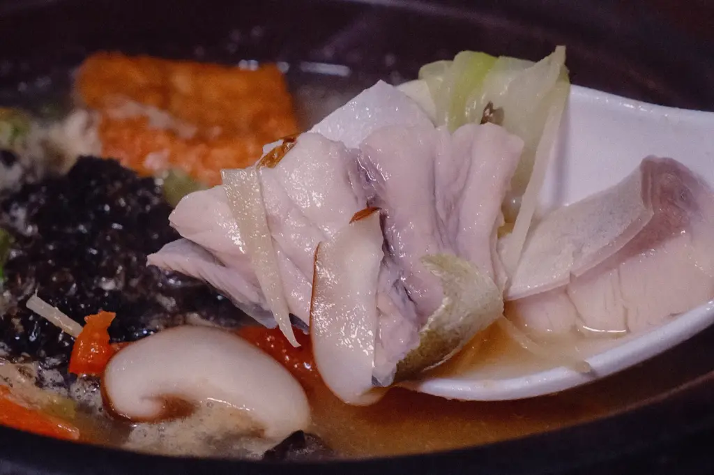 Close-up side-angle shot of tender pomfret fish lifted from a clear collagen fish broth with shiitake mushroom, vegetables, and aromatic seasonings in a bowl of traditional Singapore pomfret fish soup.