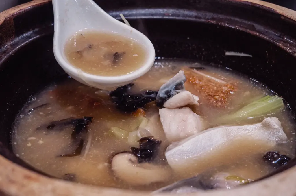 High-angle close-up shot of a white spoon gathering rich fish broth above a claypot filled with pomfret slices, mushrooms, and cabbage.