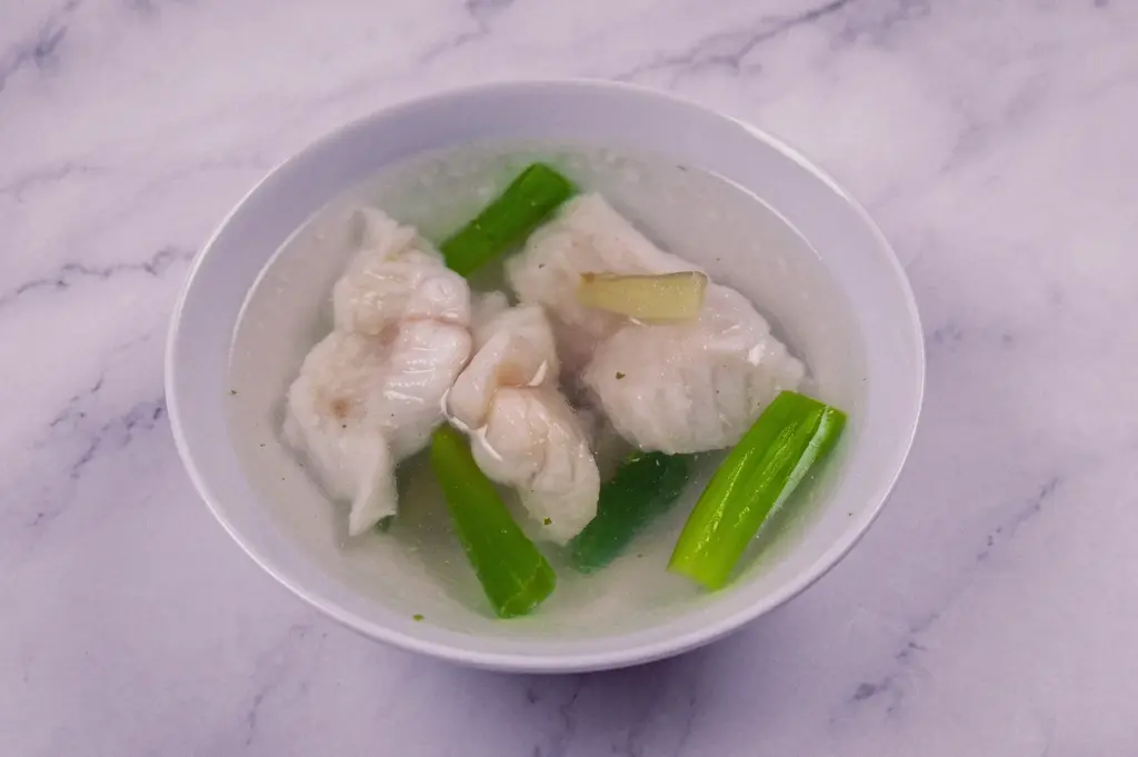 Top-down overhead shot of a light, clea fish soup served in a white bowl on a marble countertop, containing delicate poached white fish fillets, sliced ginger, and fresh green spring onion segments in a translucent broth.