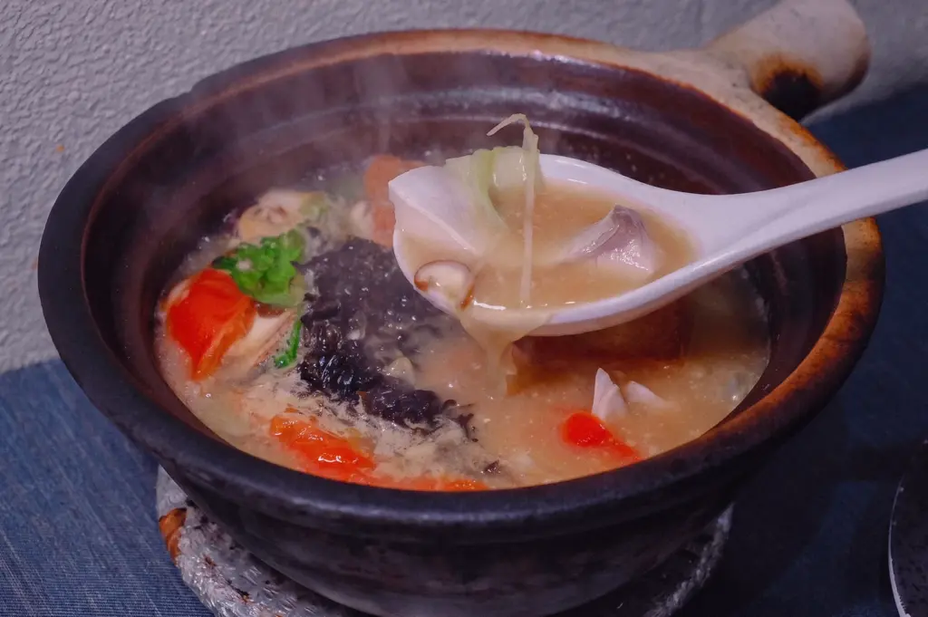 Close-up side-angle shot of pomfret fish soup in a claypot with a spoon lifting rich collagen broth, fish slices, vegetables, shiitake mushroom, and tomato, highlighting a generous restaurant fish soup portion.