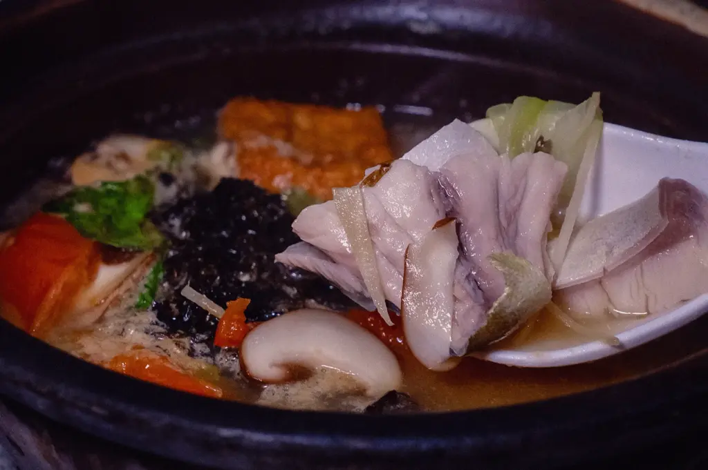 Extreme close-up shot of a white spoon holding a tender slice of fresh pomfret fish over a claypot of Hup Lok's Grandpa's Pomfret Fish Soup, garnished with sliced shiitake mushrooms, tomatoes, ginger, and sea vegetable ingredients.