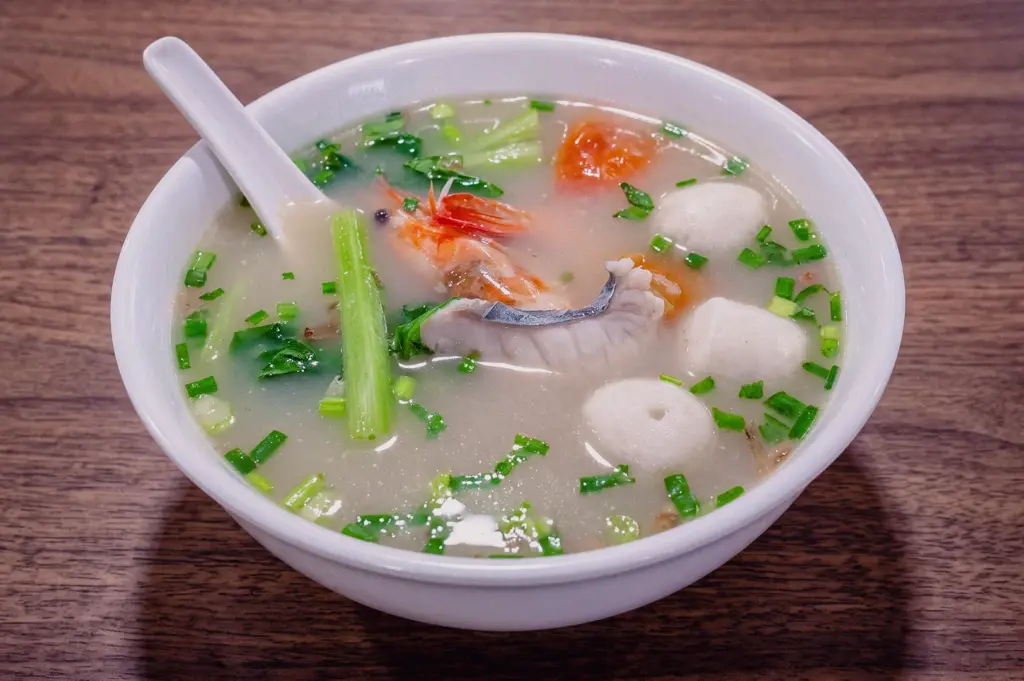 Bowl of Singapore seafood fish soup with fresh prawns, fish slices, fish balls and vegetables in clear broth, photographed from a 45-degree overhead angle on a wooden table.