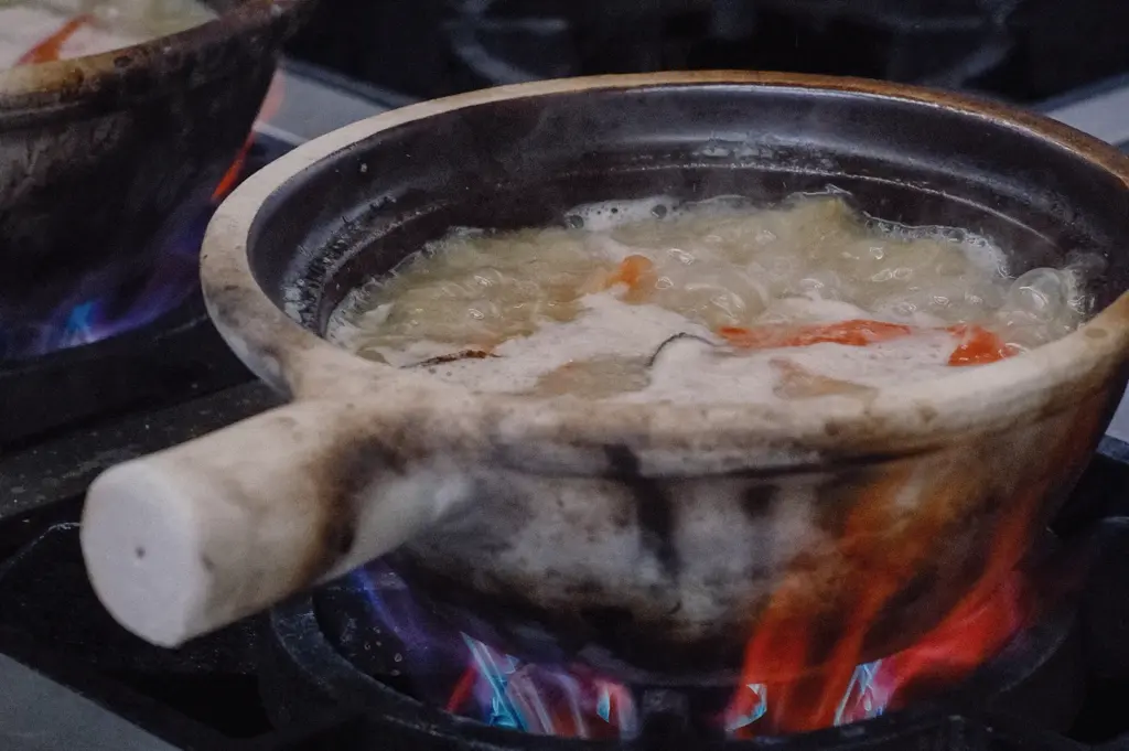 Eye-level close-up of collagen-rich fish broth simmering in a traditional claypot over an open flame, showcasing the slow-cooking process behind Singapore fish soup.