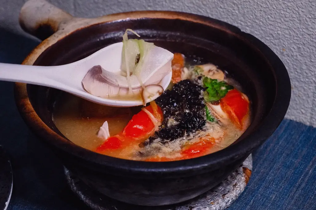 Eye-level close-up shot of a white spoon scooping a tender piece of pomfret from a steaming claypot of rich broth studded with tomatoes, seaweed, and ginger.