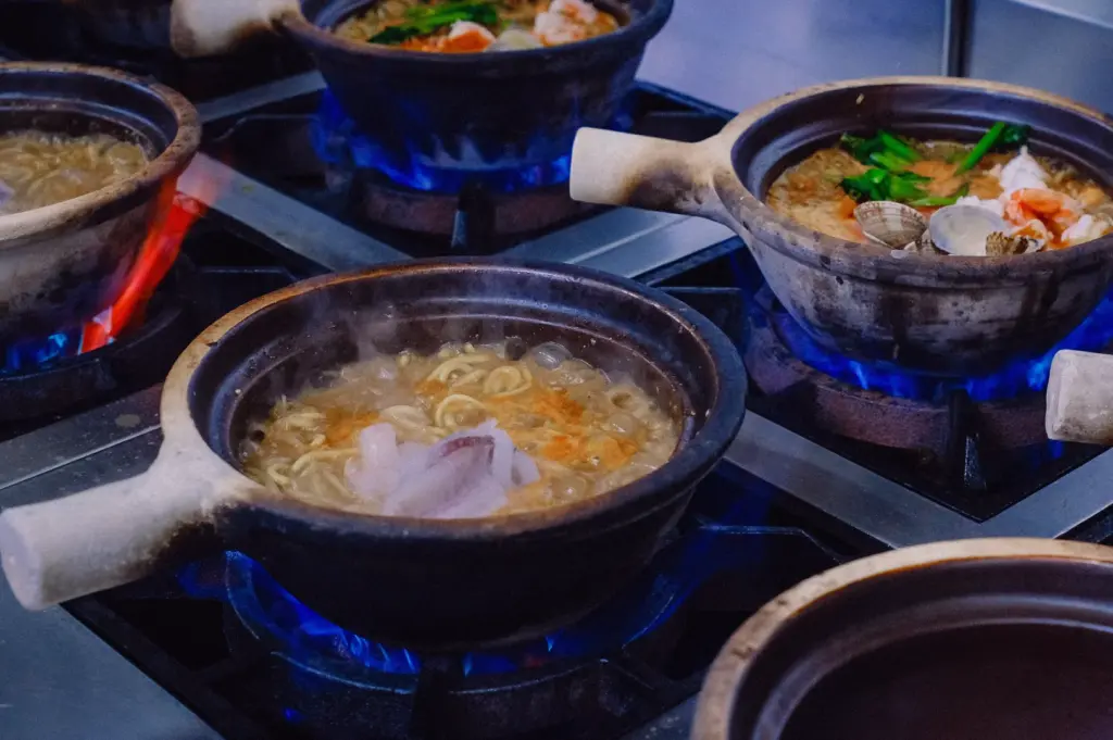 High-angle view of multiple clay pots simmering restaurant fish soup over blue gas flames, featuring collagen-rich fish broth, seafood, vegetables and noodles in a professional Singapore kitchen.