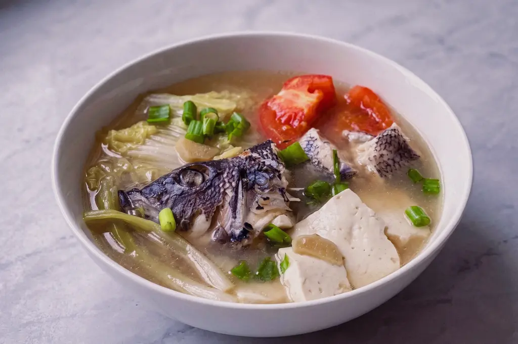 Eye-level view of collagen-rich fish head soup with tofu, tomatoes, napa cabbage and savoury fish bone broth, highlighting the rich texture and deep flavour of traditional fish head soup.