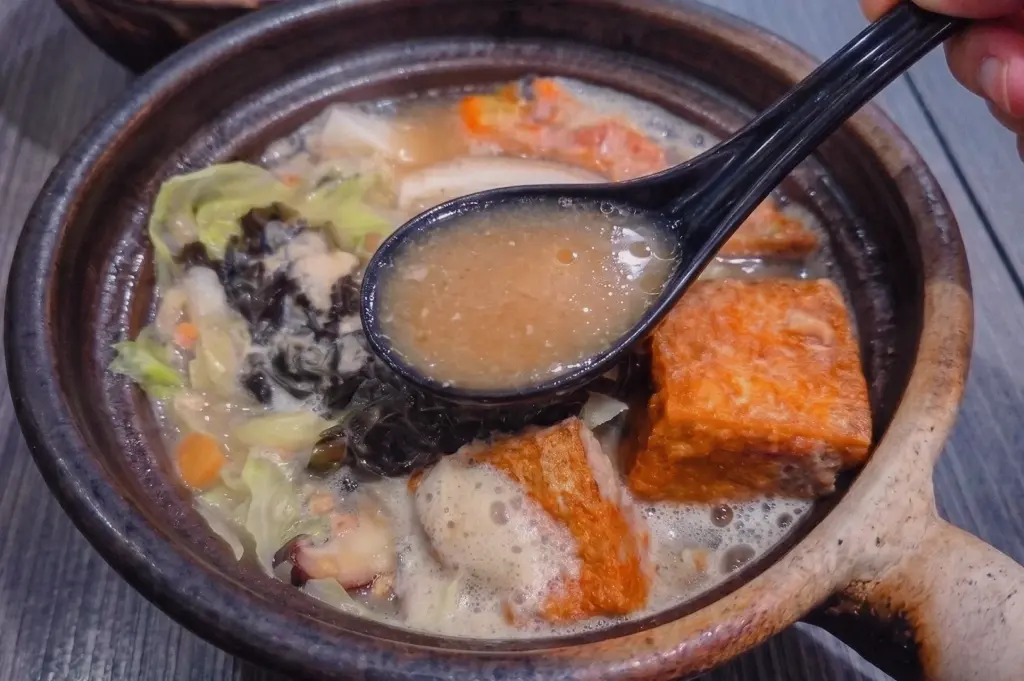 Close-up angle of a spoon lifting collagen-rich pomfret fish soup broth from a traditional claypot, showcasing the savoury homemade fish stock with tofu, cabbage, shiitake mushrooms, and seaweed.
