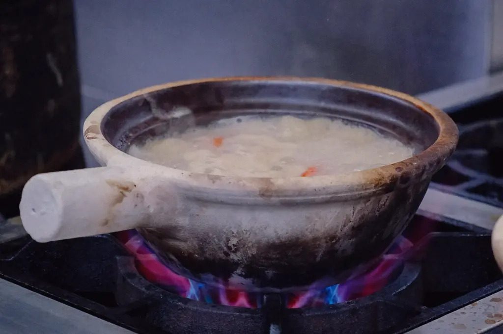 Eye-level side view of collagen-rich fish broth simmering in a traditional claypot over an open flame, showcasing the preparation of Singapore-style pomfret fish soup.