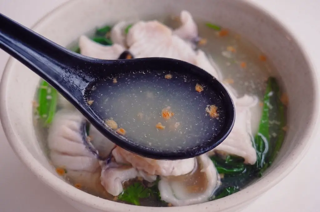 Close-up, high-angle shot of a black spoon scooping the clear, collagen-rich fish soup broth seasoned with fried garlic bits from a bowl filled with fish slices.