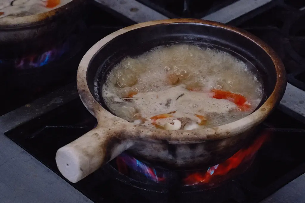 High-angle medium shot showing a single claypot of pomfret fish soup actively bubbling on a gas stove flame in a kitchen setup.