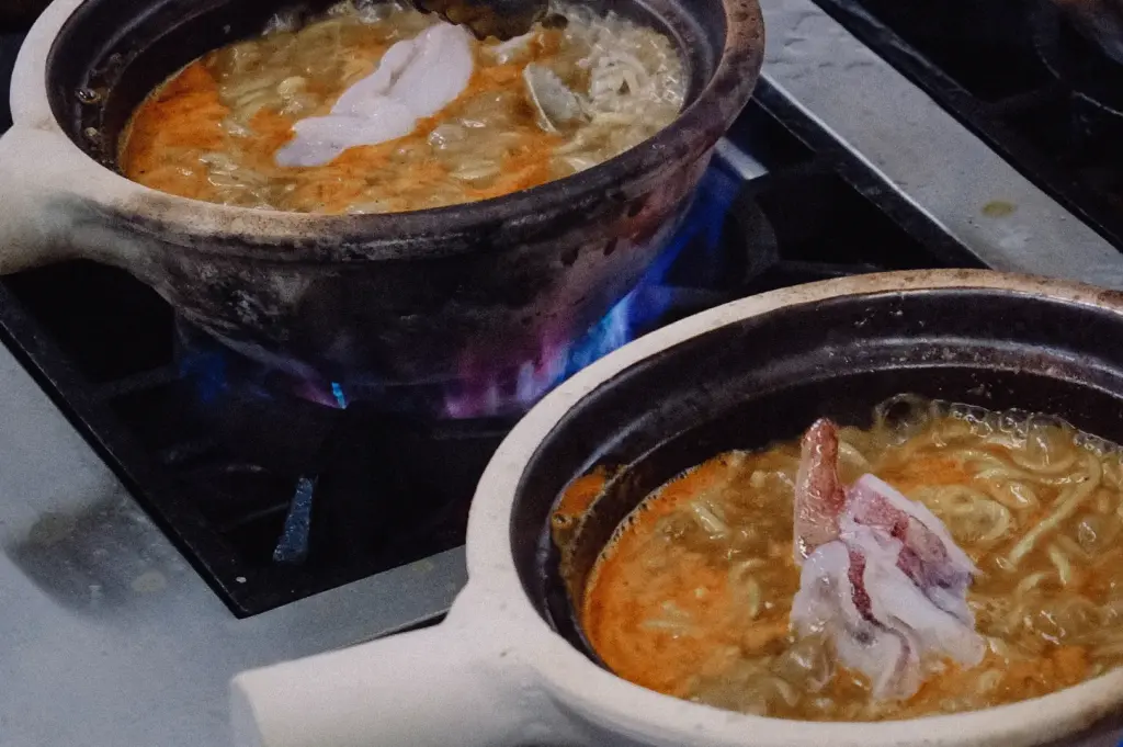 High-angle medium shot showing two rustic claypots simmering on gas burners with fresh pomfret slices added to the hot broth.