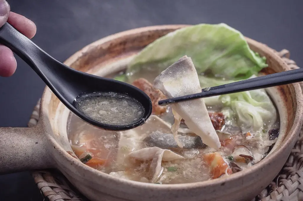 Eye-level close-up shot of chopsticks lifting a tender pomfret fish slice next to a black spoon holding rich soup over a claypot.