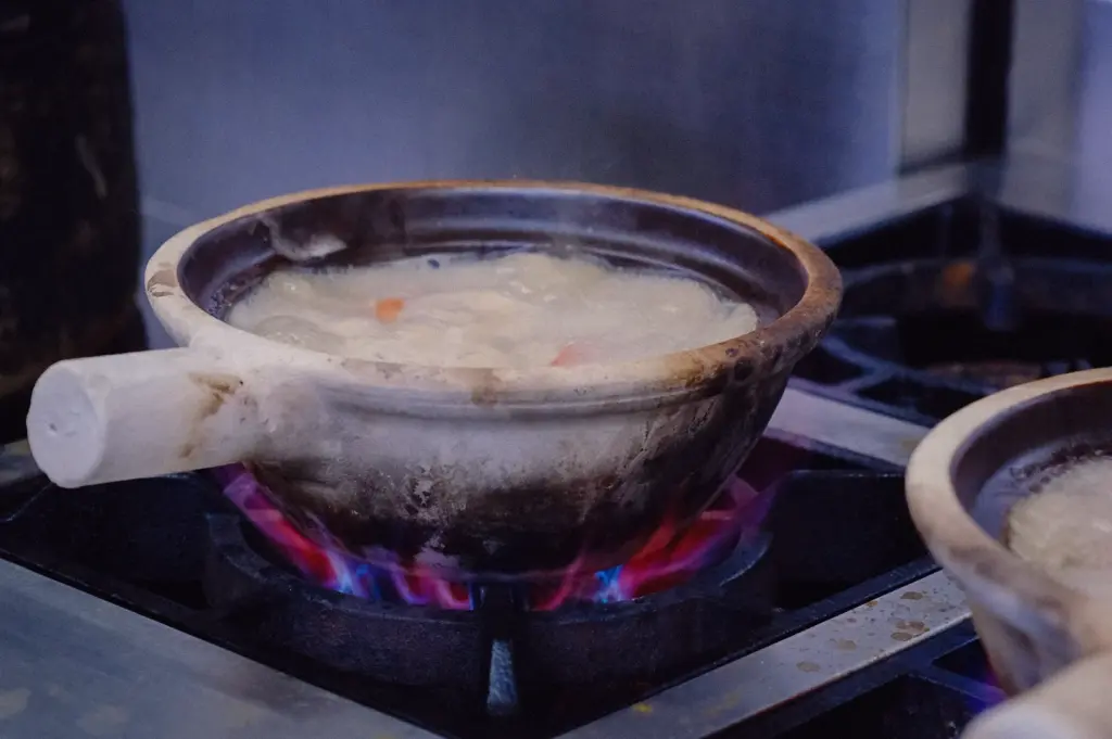 Eye-level close-up shot of a traditional claypot filled with hot, bubbling fish soup broth simmering directly over a blue gas stove flame in a kitchen.