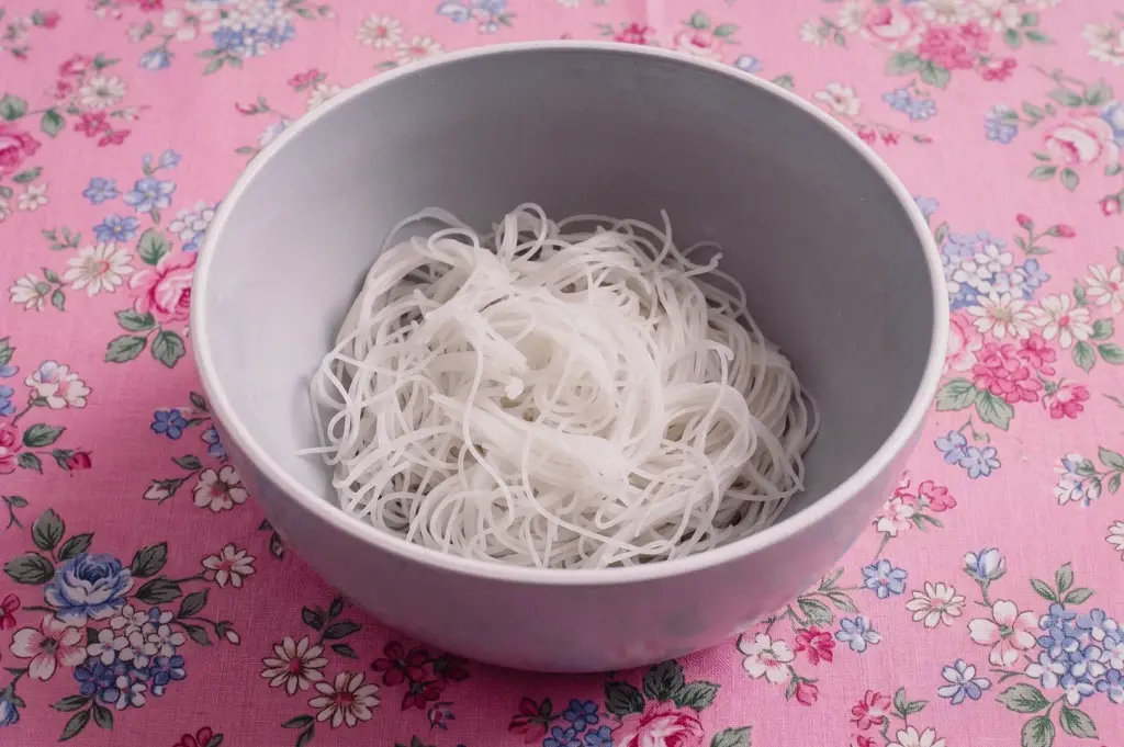 White bowl containing a small portion of thin white rice vermicelli (bee hoon) sits on a pink floral-patterned tablecloth. The noodles are loosely arranged in the center of the bowl, while the tablecloth features scattered pink, blue, and white flower designs. Soft natural light illuminates the scene, highlighting the delicate texture of the noodles and the pastel colors of the background.