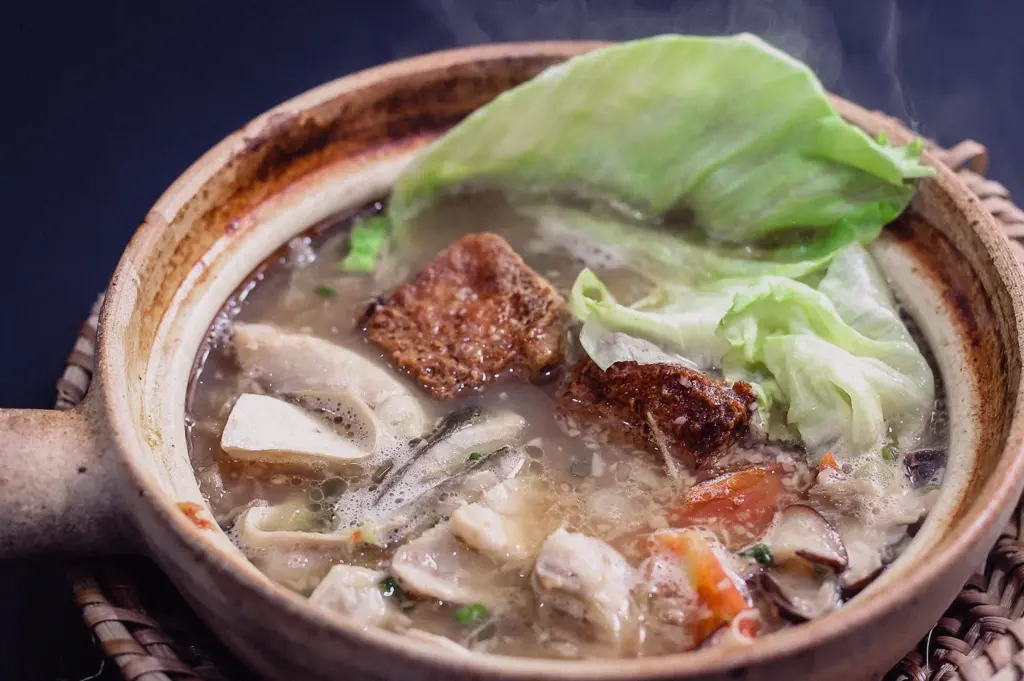 High-angle, close-up shot of a steaming hot, bubbling claypot packed with simmered Singapore fish soup, fresh fish slices, green lettuce leaves, tomatoes, and seaweed in a thick, rich collagen-based broth.