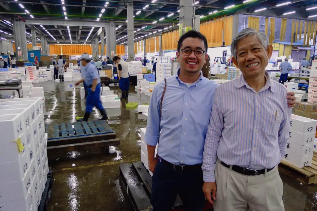 Eye-level wide-angle portrait shot taken at Tsukiji Market in Tokyo, showing seafood buyers inside a bustling wholesale fish market surrounded by fresh seafood vendors and fish boxes, highlighting the sourcing expertise behind premium Singapore fish soup.
