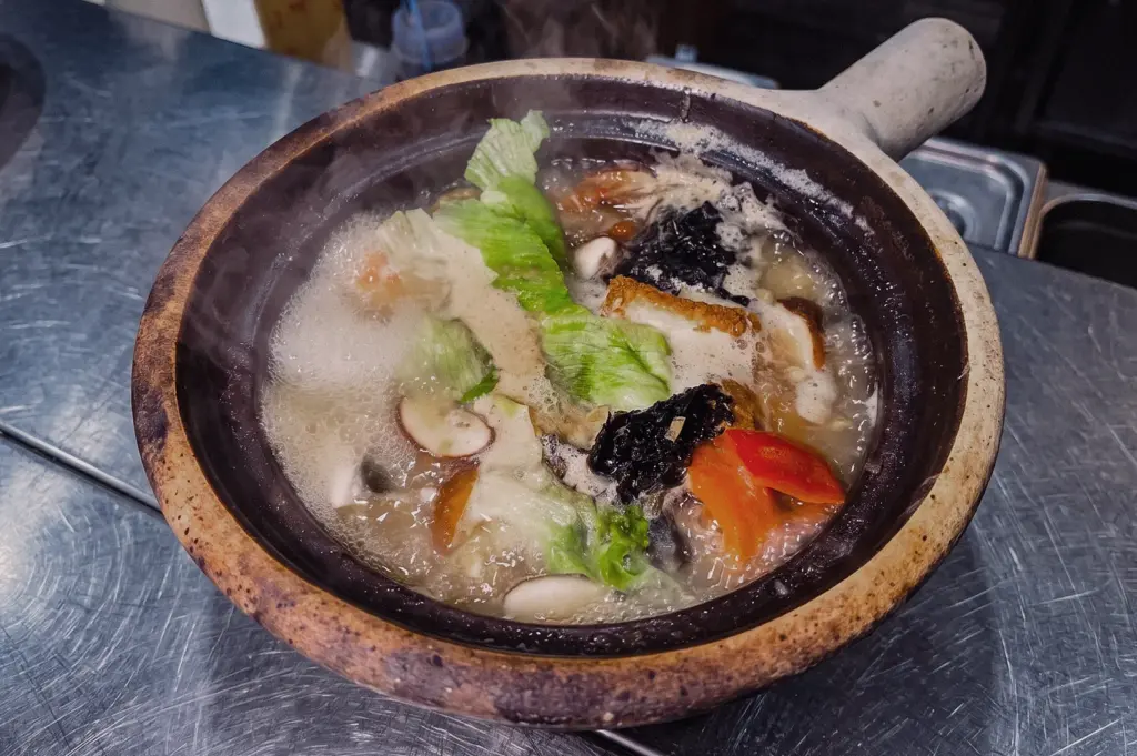 High-angle three-quarter view of Grandpa's Pomfret Fish Soup served in a claypot with pomfret fish, shiitake mushrooms, lettuce, tomato and collagen-rich broth.