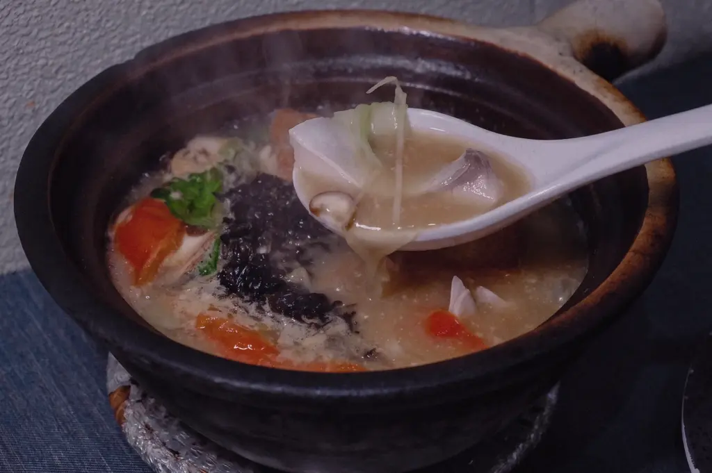 A close-up food photography image captured from a slight side angle showing a steaming claypot fish soup. A white ceramic spoon lifts thick collagen-rich broth along with cabbage and mushrooms from the claypot, while tomatoes, leafy greens and fish ingredients remain visible in the bubbling soup below.