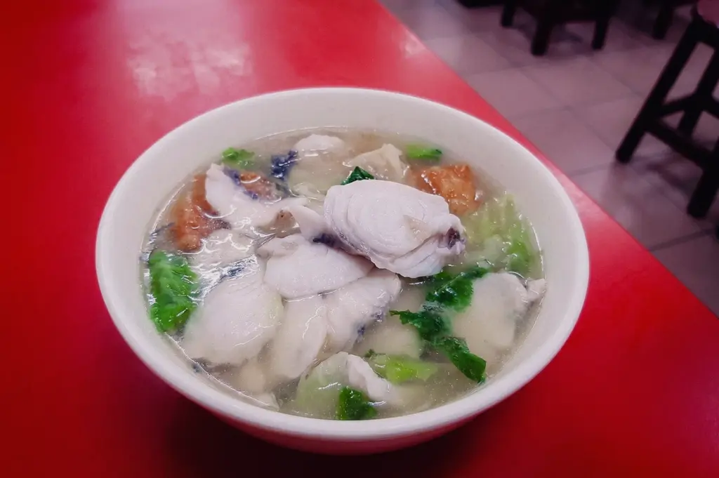 Eye-level three-quarter angle shot of traditional hawker fish soup in a simple bowl with sliced fish, leafy vegetables and clear broth, served at a Singapore coffee shop for an affordable everyday meal.